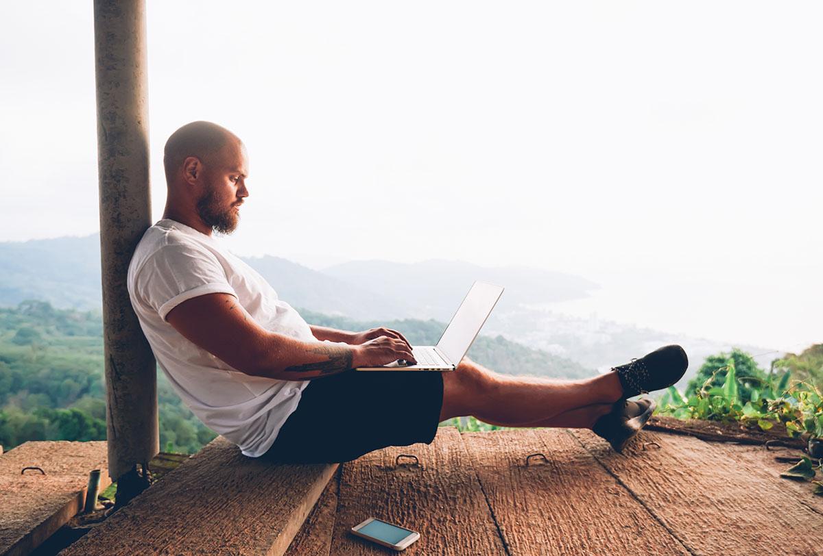 A man seated on a wooden bench, focused on his laptop, surrounded by a serene outdoor environment.