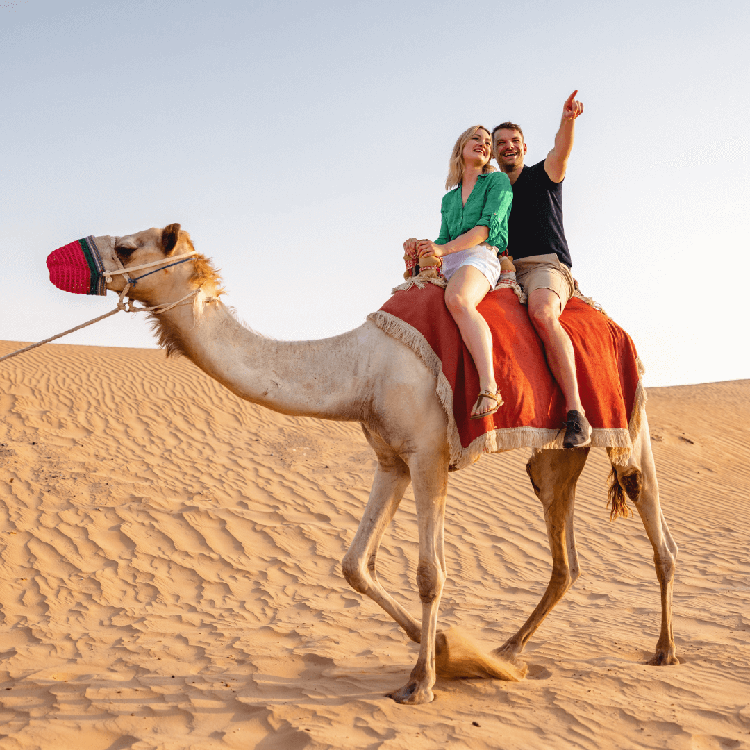 A couple riding a camel in the desert, surrounded by sand dunes under a sunny sky.