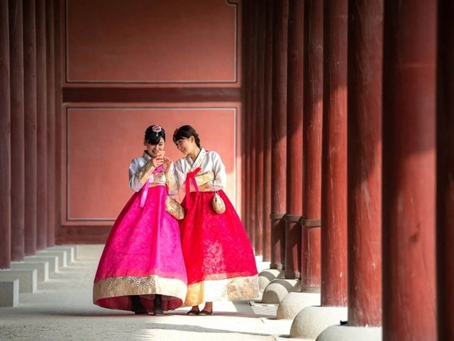 Two individuals wearing traditional Korean hanboks stand in a corridor with wooden pillars and red walls. The setting suggests a historical or cultural significance, complementing their attire.