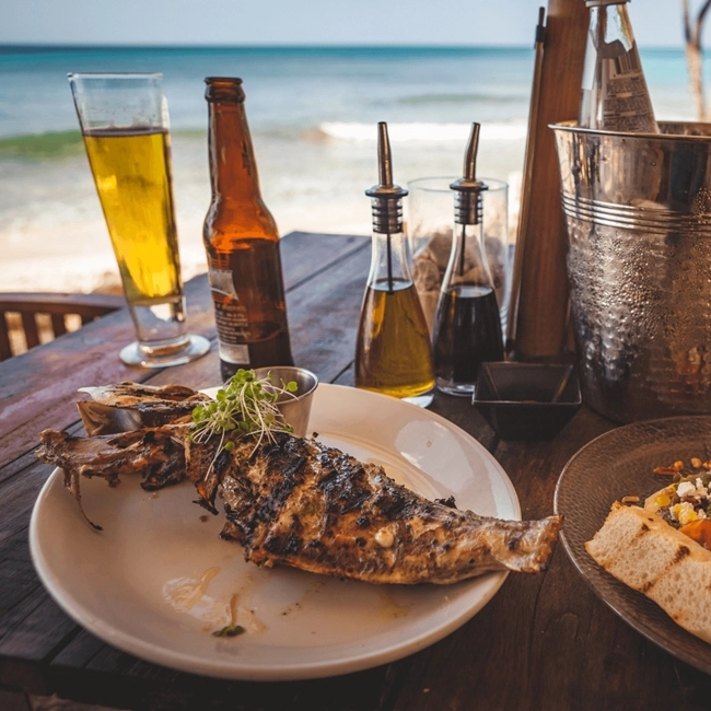 The image shows a meal of grilled fish on a plate, with condiments and a glass of beer on a table overlooking the beach. It highlights a relaxed dining experience by the sea.