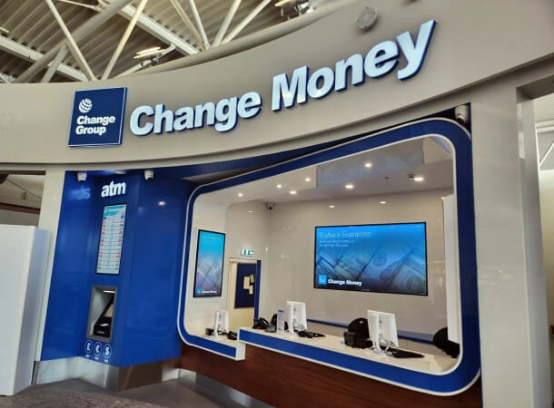 The image displays a currency exchange counter labeled “Change Money” with an ATM on the side. It likely serves as a financial service point, possibly located in an airport or similar transit area. The presence of multiple currencies displayed on