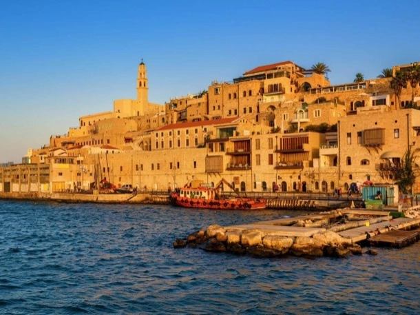 The image depicts a coastal scene at sunset with buildings of varying heights, some with terracotta roofs, along a waterfront promenade. A church tower rises above the other structures, and there is a boat docked near the stone pier, all bathed in warm light. This image is interesting for its picturesque representation of Mediterranean architecture and the serene maritime atmosphere it conveys