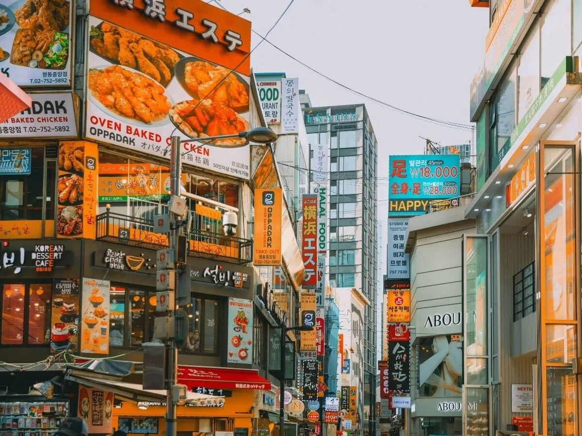 The image shows a vibrant street scene with numerous signs in various languages, including English and Korean. The signs advertise a variety of food options such as baked chicken, oven-baked ribs, and other dishes. The architecture suggests an urban environment, possibly within a shopping or dining district. This image is interesting due to the cultural blend it represents and the bustling atmosphere it suggests.