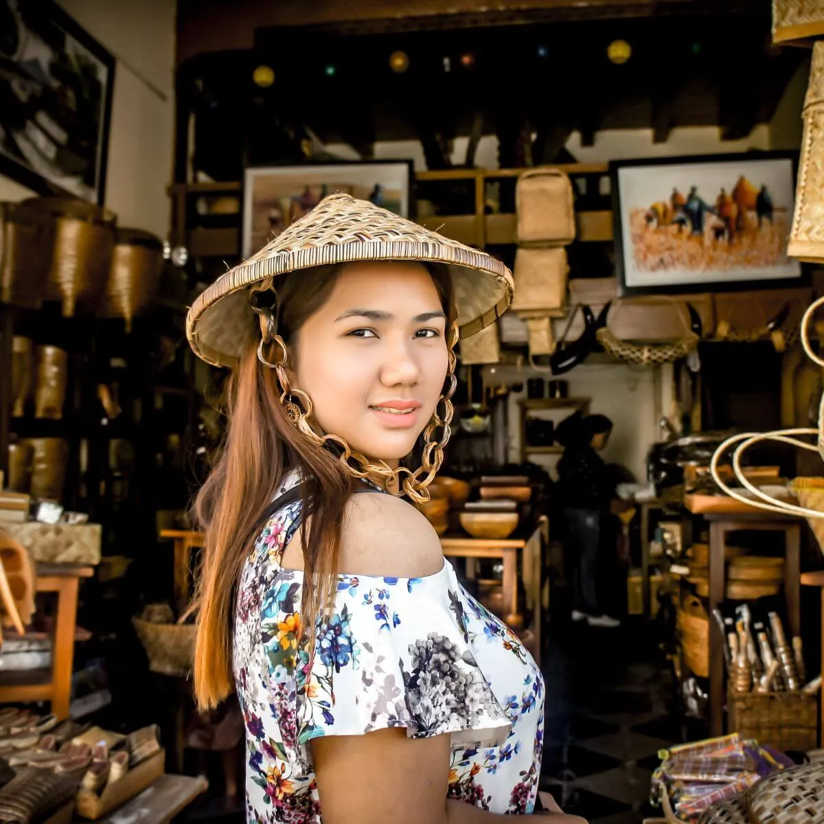 A woman wearing a hat stands in a shop, surrounded by various items for sale.