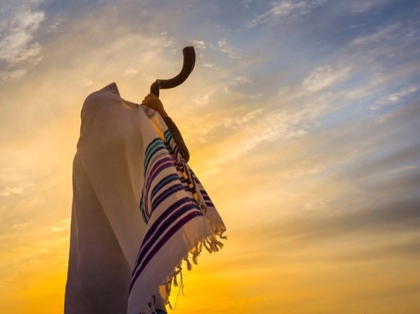 The image shows a shofar, a traditional Jewish horn, placed against a draped tallit, or prayer shawl, with the backdrop of a sunrise or sunset sky. This scene is significant as it represents elements of Jewish tradition and religious practice.