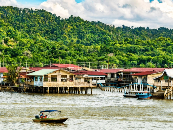 The image displays a waterfront village with stilt houses extending over the water, backed by a lush green hill, showcasing a lifestyle intertwined with nature. A small boat is in the foreground, indicating the primary mode of transportation in this community. 