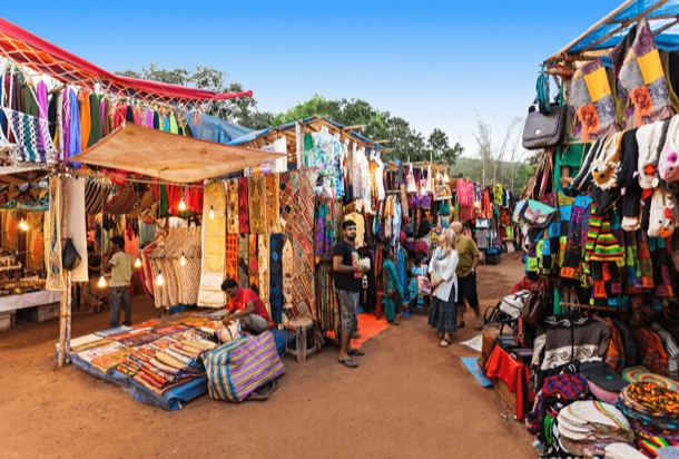 The image shows a vibrant outdoor market with various stalls displaying colorful textiles, clothing, and crafts. Shoppers are browsing the items, highlighting the market’s role as a hub of commerce and cultural exchange.