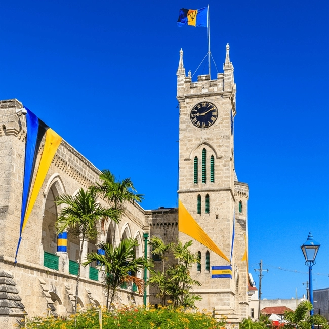 The image shows a historic stone building with a clock tower and flying flags under clear blue skies. The architectural style and national flags suggest its cultural significance.