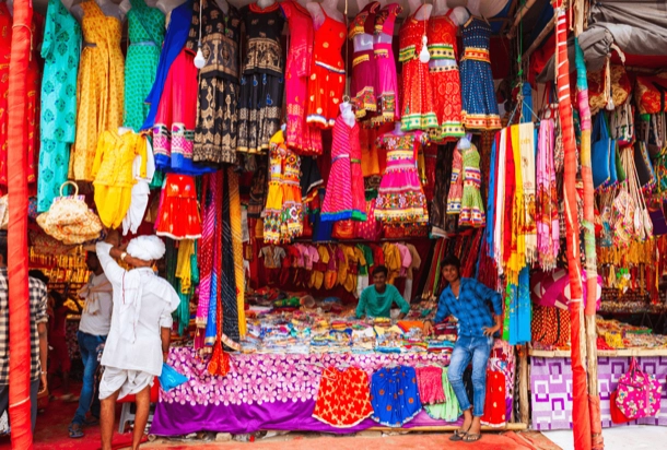 The image displays a vibrant and colorful street market stall with an array of traditional clothing and accessories. It is interesting due to the rich variety of patterns and hues, showcasing cultural fashion