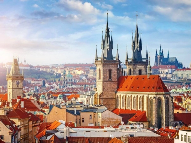 The image shows a panoramic view of a historic cityscape with Gothic architecture, including spires and a prominent church under a clear sky. The scene is interesting for its blend of medieval structures and modern urban life.