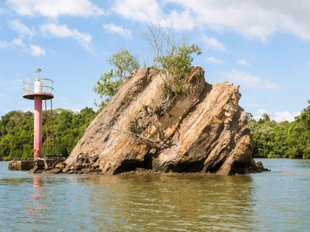 The image displays a large rock formation emerging from a body of water, with a small lighthouse structure to the left. The scene is set against a backdrop of blue sky and lush greenery, making it an interesting blend of natural and man-made elements. 