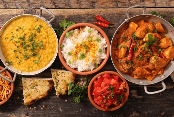 The image displays a variety of dishes on a wooden surface, including what appears to be lentil curry, rice with herbs, chicken curry, garlic bread, and a tomato-based side dish. This array of food is interesting as it suggests a rich culinary diversity, possibly indicative of Indian cuisine