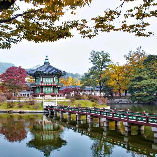 The image shows a traditional Korean-style building with a multi-tiered, ornate roof beside a calm body of water. A bridge with red railings leads to the building, framed by trees with autumn-colored leaves.