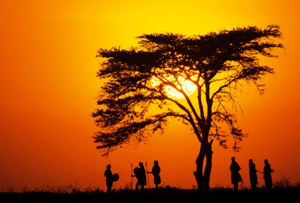 The image shows a silhouette of several individuals and a tree against an orange-hued sky, suggesting it is either sunrise or sunset. The striking contrast between the dark figures and the vibrant background evokes a sense of tranquility and natural beauty.