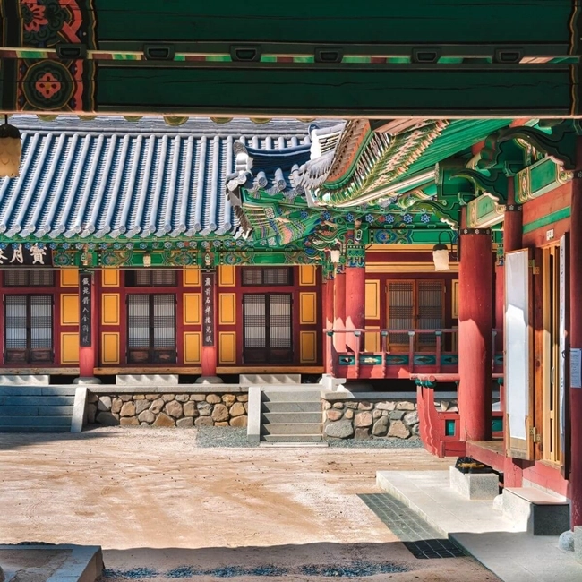 The image shows a traditional Korean Hanok with vibrant colors. The architecture features a tiled roof with intricate designs, wooden beams, and brightly painted walls in red and green, highlighting the cultural heritage of Korea.