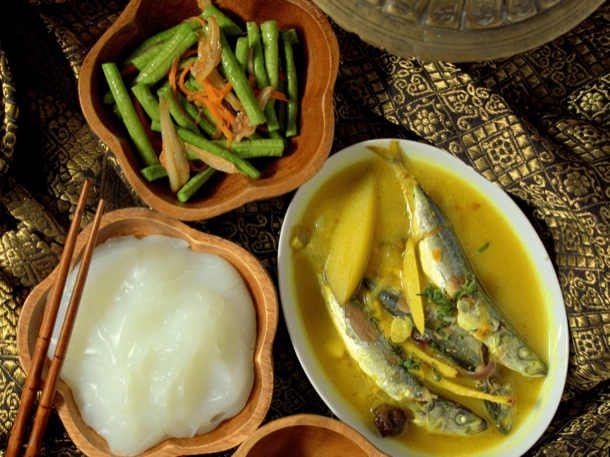 The image displays a traditional meal setup with a bowl of rice noodles, a plate containing fish in yellow curry, and a wooden bowl with green beans and carrots. The arrangement on a patterned textile suggests cultural significance, possibly showcasing cuisine from Southeast Asia. 